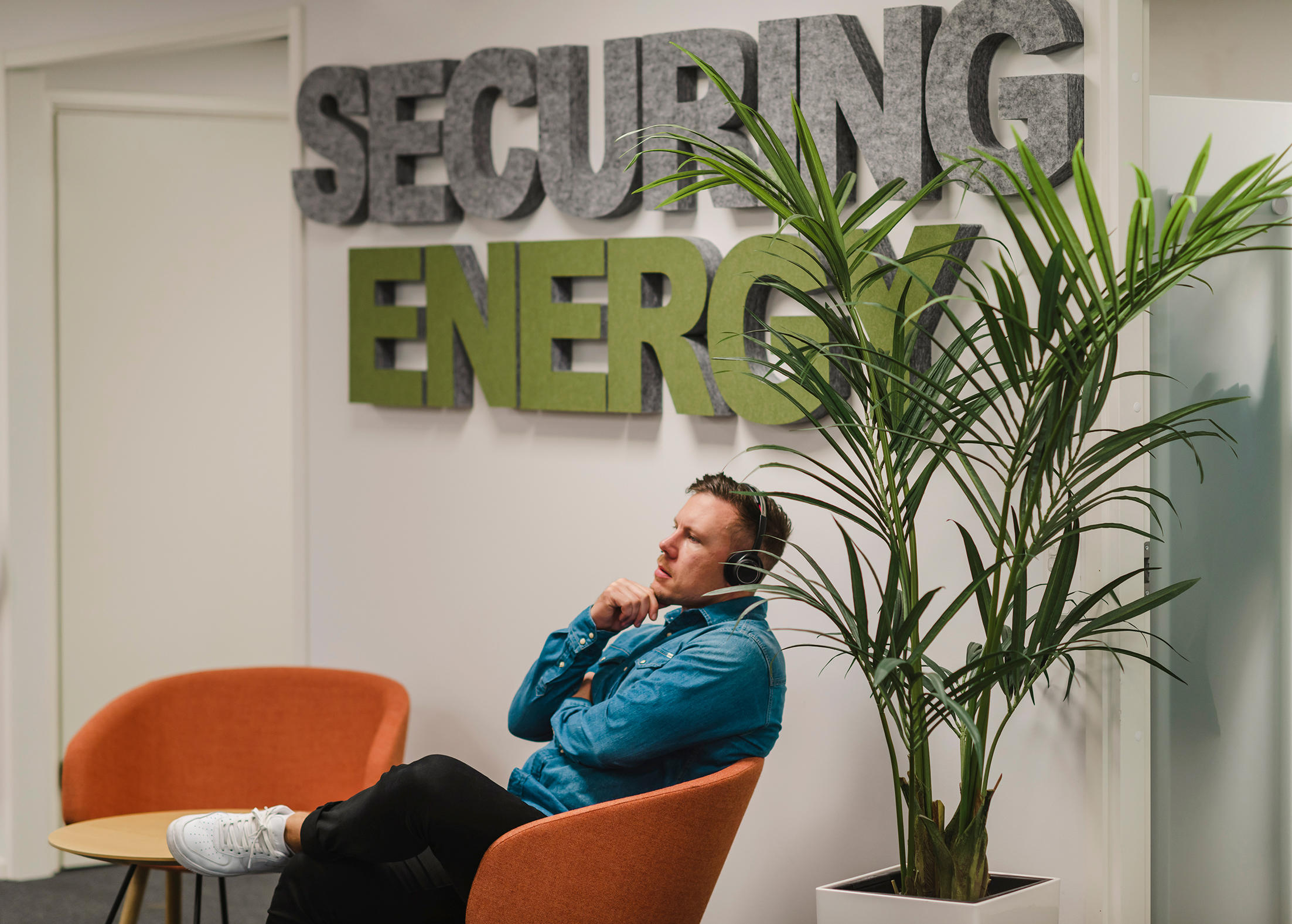 A person sitting in an orange chair in an office area. A sign behind them reads "SECURING ENERGY," and a tall potted plant is nearby.