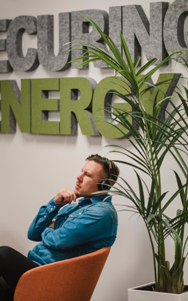 En person sitter i en orange stol i ett kontorsutrymme. En skylt bakom visar texten "SECURING ENERGY," och en hög krukväxt står i närheten.