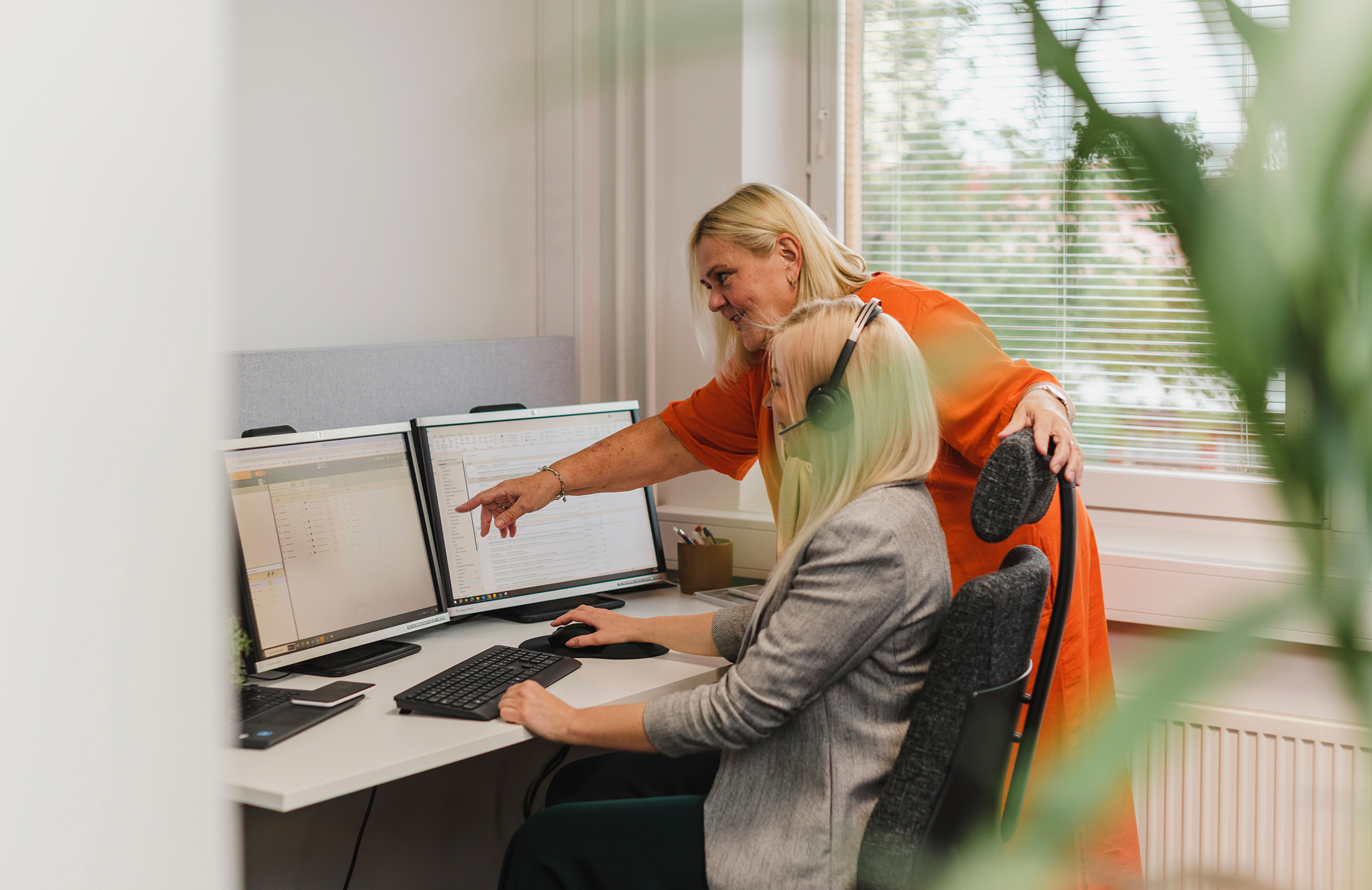 Two people in an office; one seated with headphones at a desk with dual monitors, the other standing and pointing at a screen.