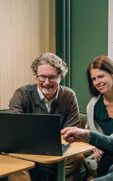 Four people sitting around a table, looking at a laptop, with a bamboo wallpaper and window in the background.