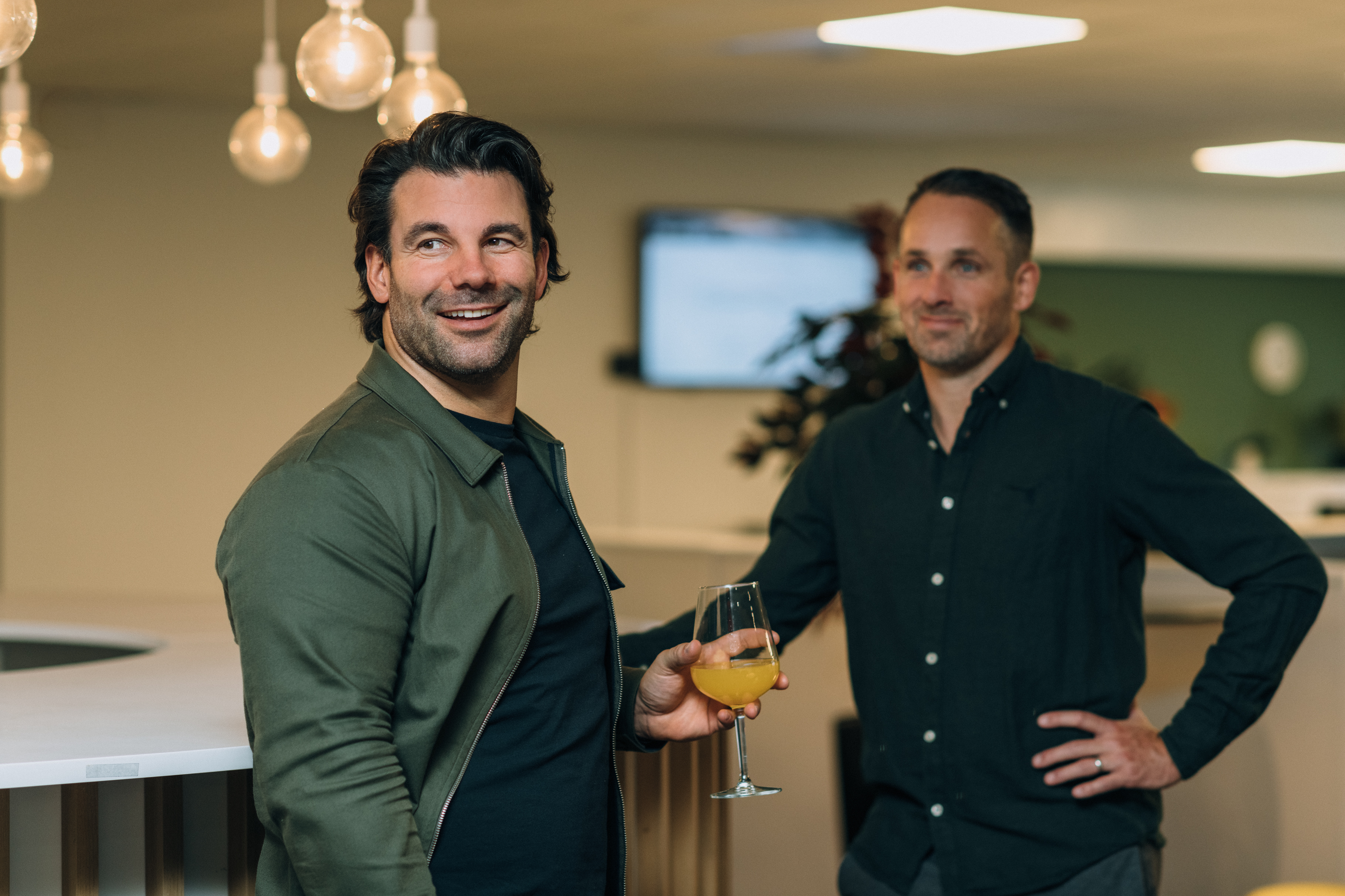 Two people by a bartop indoors, one holding a glass with orange drink, under hanging lights with a screen and plants in the background.