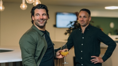 Två personer vid en bardisk inomhus, en håller ett glas med orange dryck, under hängande lampor med en skärm och växter i bakgrunden.