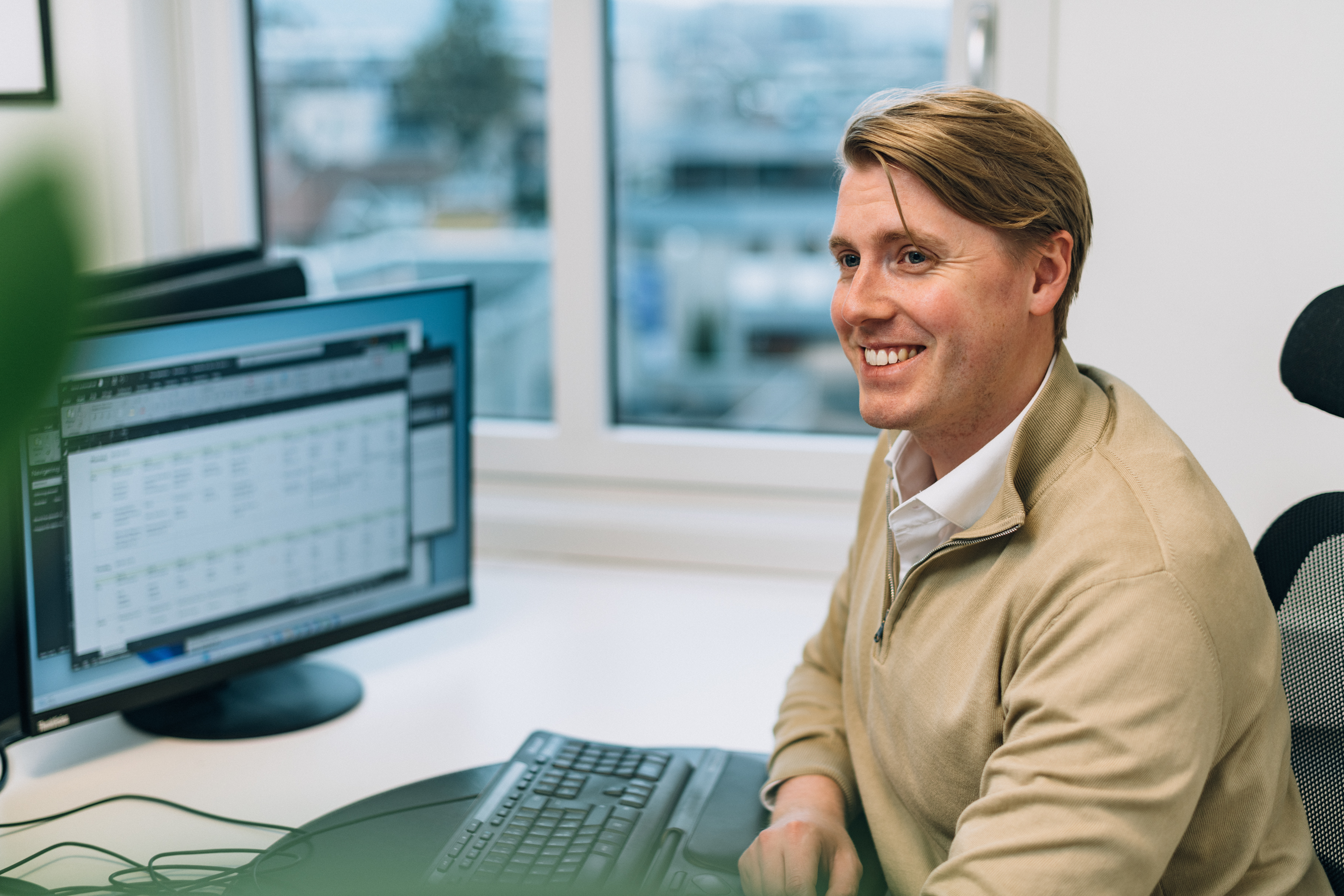 En person sitter vid ett skrivbord framför en datorskärm som visar ett kalkylblad eller en datatabell. Ett fönster bakom visar byggnader och träd utanför.