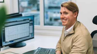 En person sitter vid ett skrivbord framför en datorskärm som visar ett kalkylblad eller en datatabell. Ett fönster bakom visar byggnader och träd utanför.