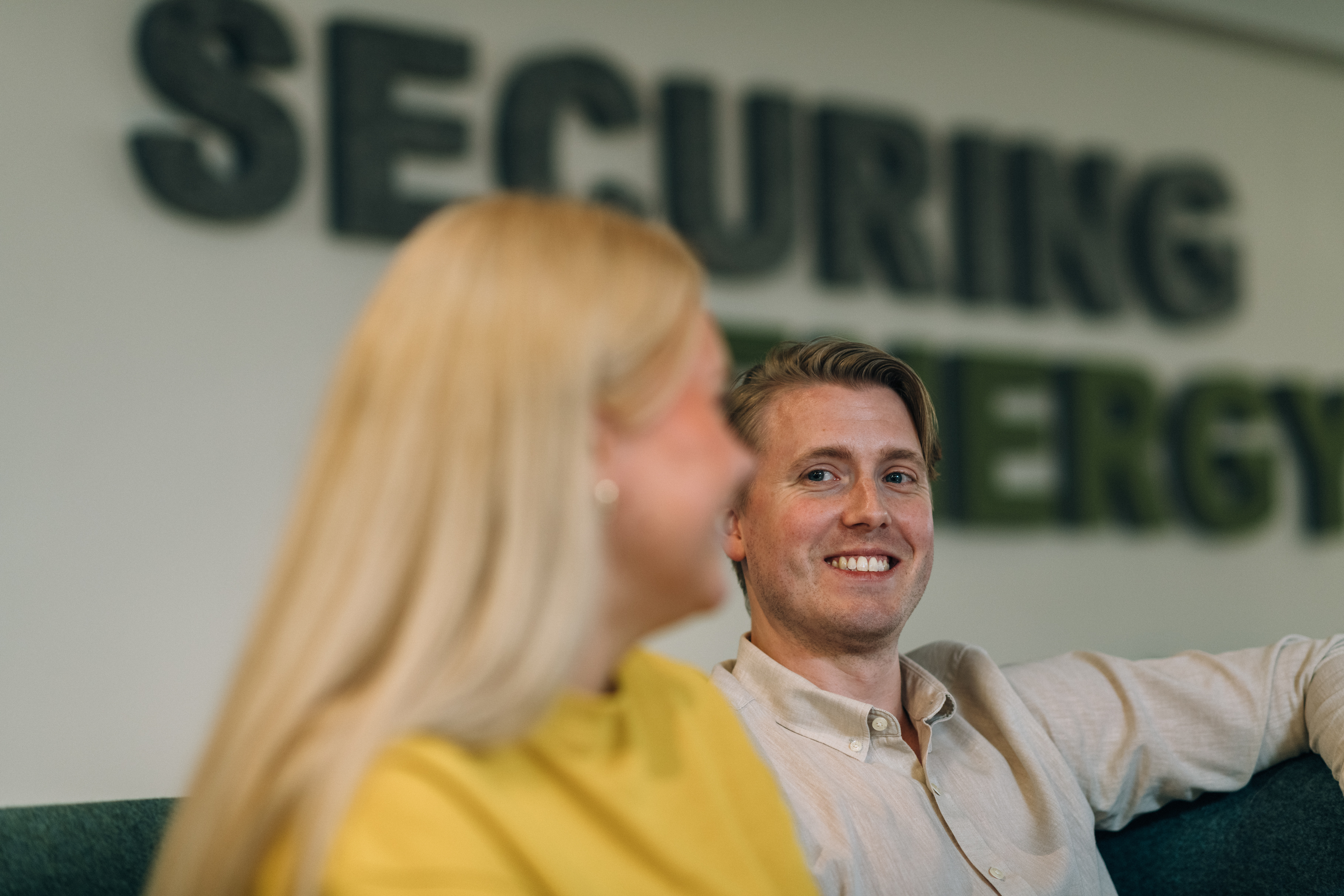 Two people sit on a couch with the text "SECURING ENERGY" visible in the background. Their faces are blurred.