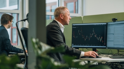 En man tittar på en skärm som visar ett finansiellt diagram med ljusstakemönster på ett kontor. En annan man arbetar vid ett skrivbord i bakgrunden.