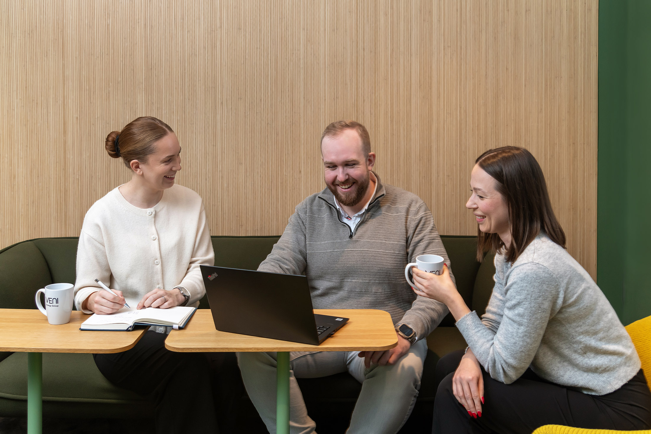 Två kvinnor och en man sitter runt ett litet träbord; en kvinna håller en penna över en anteckningsbok, mannen använder en bärbar dator och den andra kvinnan håller en mugg.