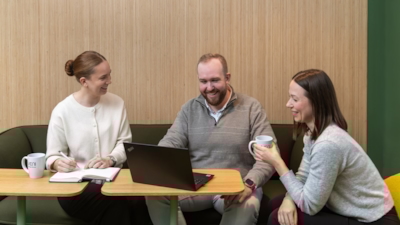 Två kvinnor och en man sitter runt ett litet träbord; en kvinna håller en penna över en anteckningsbok, mannen använder en bärbar dator och den andra kvinnan håller en mugg.