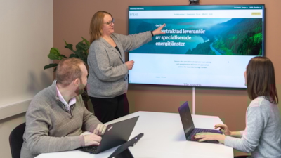 Tre personer i ett mötesrum; en står och pekar på en skärm som visar webbplatsen eneas.se, medan två andra sitter med bärbara datorer.